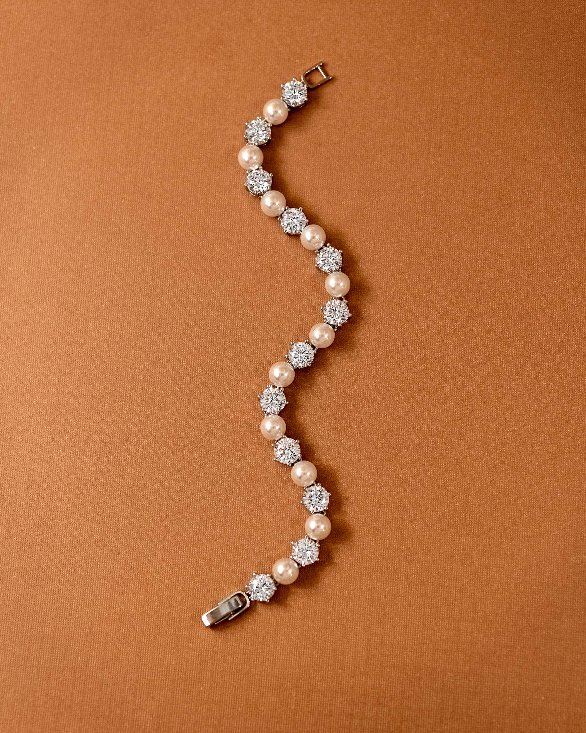 Pearl Bracelet Alternating CZ Stones A curved tennis bracelet featuring cream-colored pearls and clear round-cut gemstones in silver-toned settings, displayed on a textured brown background.