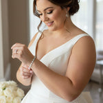 A Pearl Bracelet Alternating CZ Stones  woman in a white sleeveless wedding dress looks down at a pearl bracelet she is adjusting on her wrist, with a bouquet of white roses in the foreground and a softly lit indoor background.