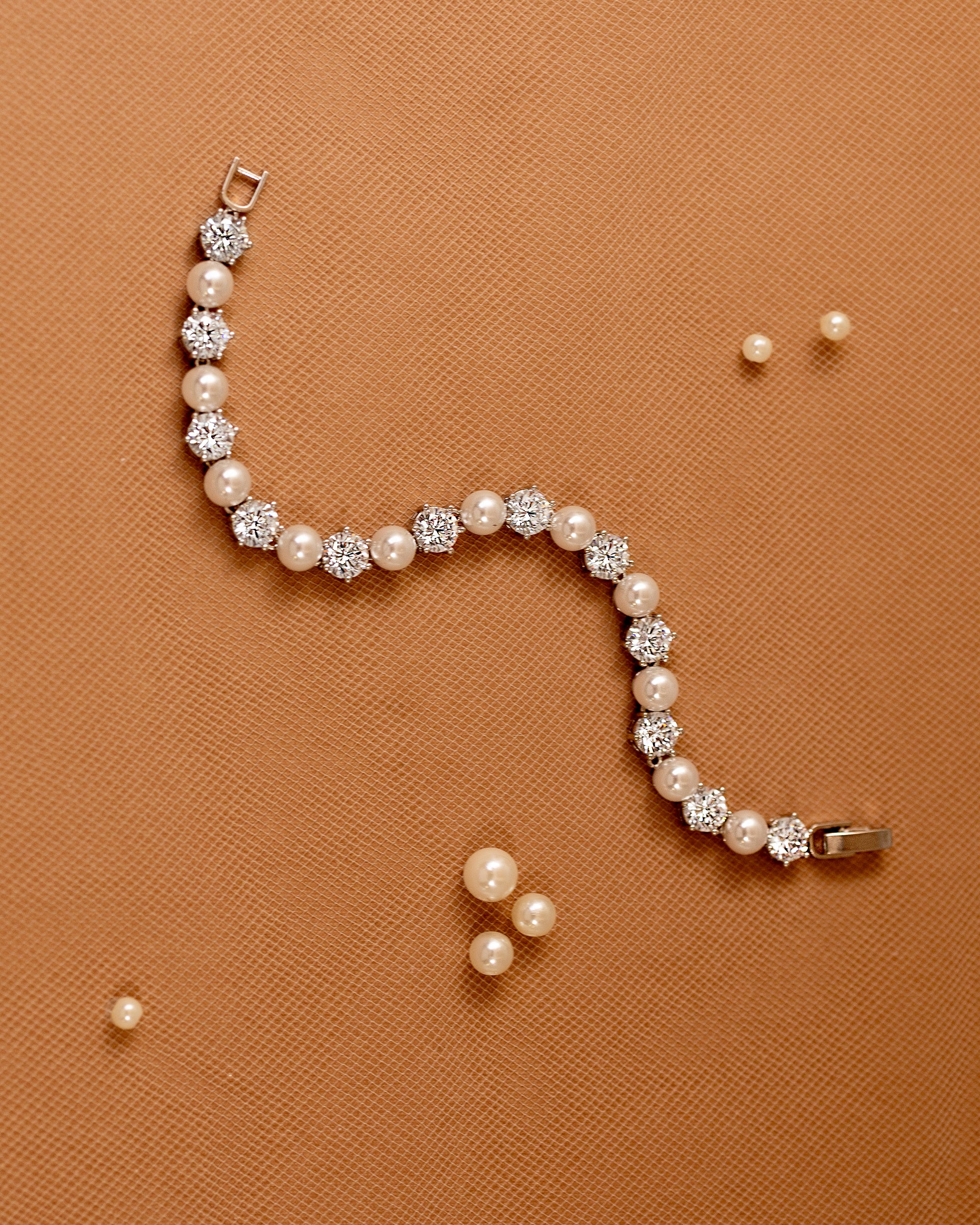 Pearl Bracelet Alternating CZ Stones A pearl and crystal bracelet arranged in a curved S-shape on a textured orange-brown background, with a few loose pearl-like beads scattered nearby.
