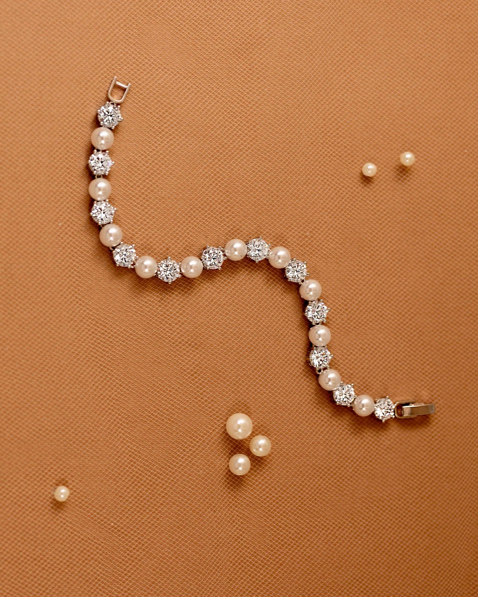 Pearl Bracelet Alternating CZ Stones A pearl and crystal bracelet arranged in a curved S-shape on a textured orange-brown background, with a few loose pearl-like beads scattered nearby.