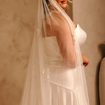 Bridal Luxe Veil with Painted Flowers Cassandra Lynne  A woman wearing a ivory wedding dress and a long sheer tulle bridal veil, posed indoors against a warm beige wall.