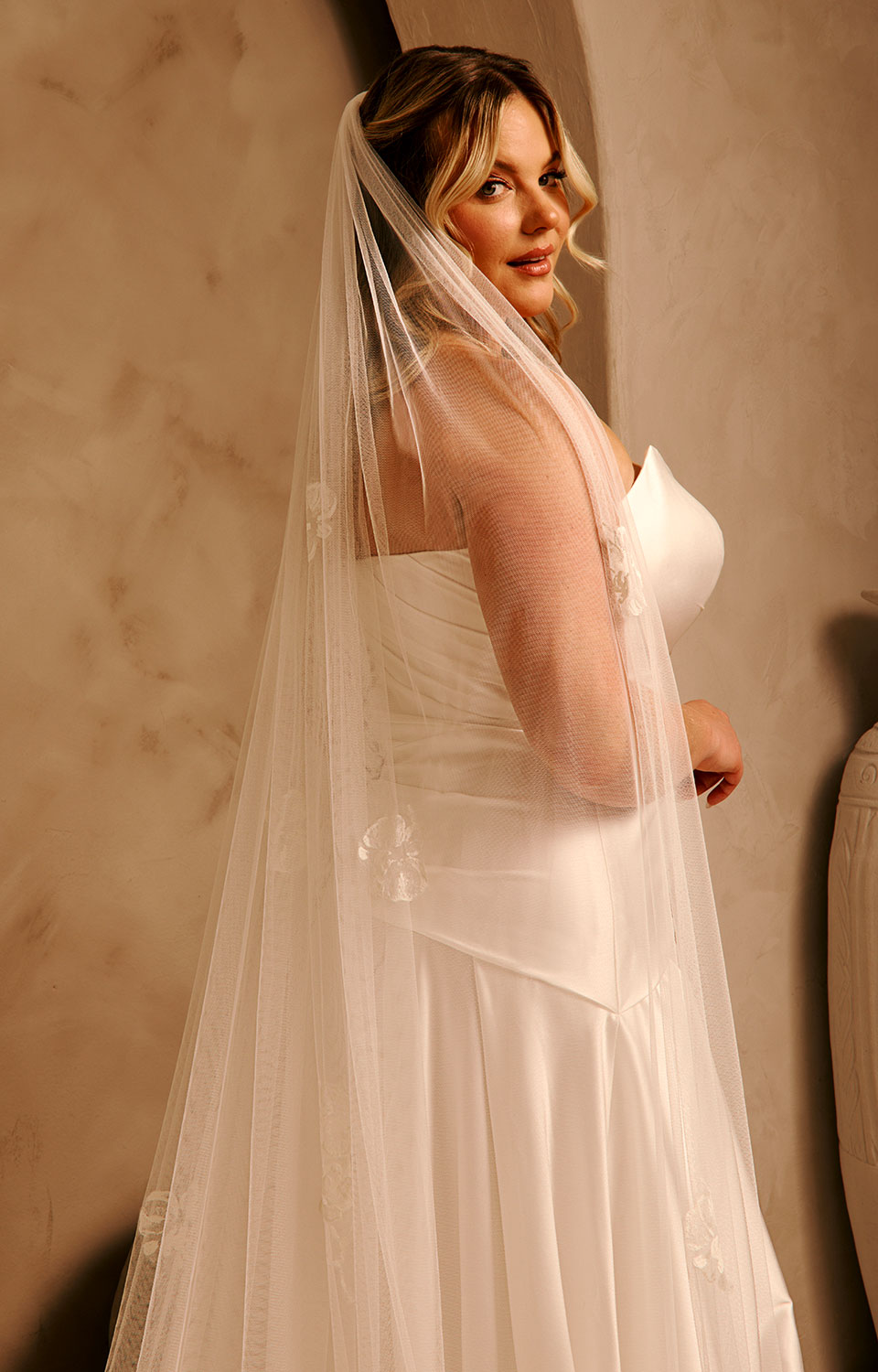 Bridal Luxe Veil with Painted Flowers Cassandra Lynne  A woman wearing a ivory wedding dress and a long sheer tulle bridal veil, posed indoors against a warm beige wall.