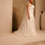 Bridal Luxe Veil with Painted Flowers Cassandra Lynne  A woman wearing a ivory wedding dress with a long sheer veil stands in a warm beige architectural setting with arches and a large vase in the background.