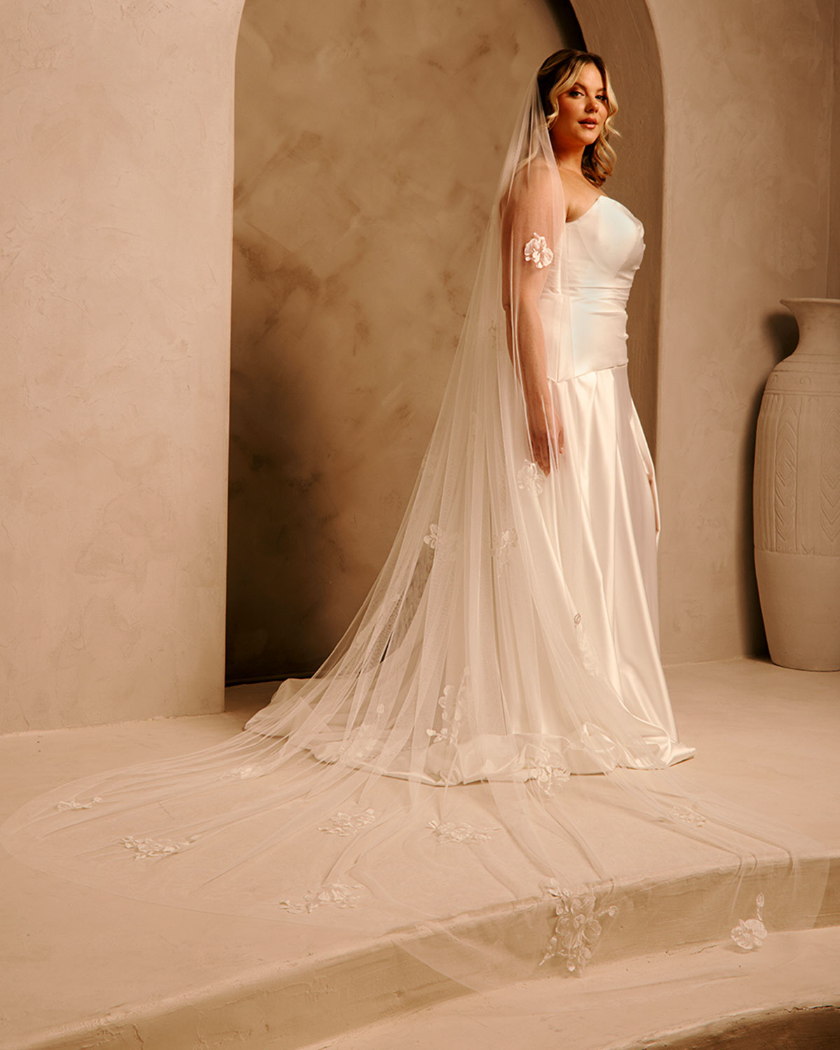 Bridal Luxe Veil with Painted Flowers Cassandra Lynne  A woman wearing a ivory wedding dress with a long sheer veil stands in a warm beige architectural setting with arches and a large vase in the background.