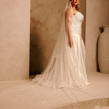 Bridal Luxe Veil with Painted Flowers Cassandra Lynne  A woman wearing a ivory wedding dress with a long sheer veil stands in a warm beige architectural setting with arches and a large vase in the background.