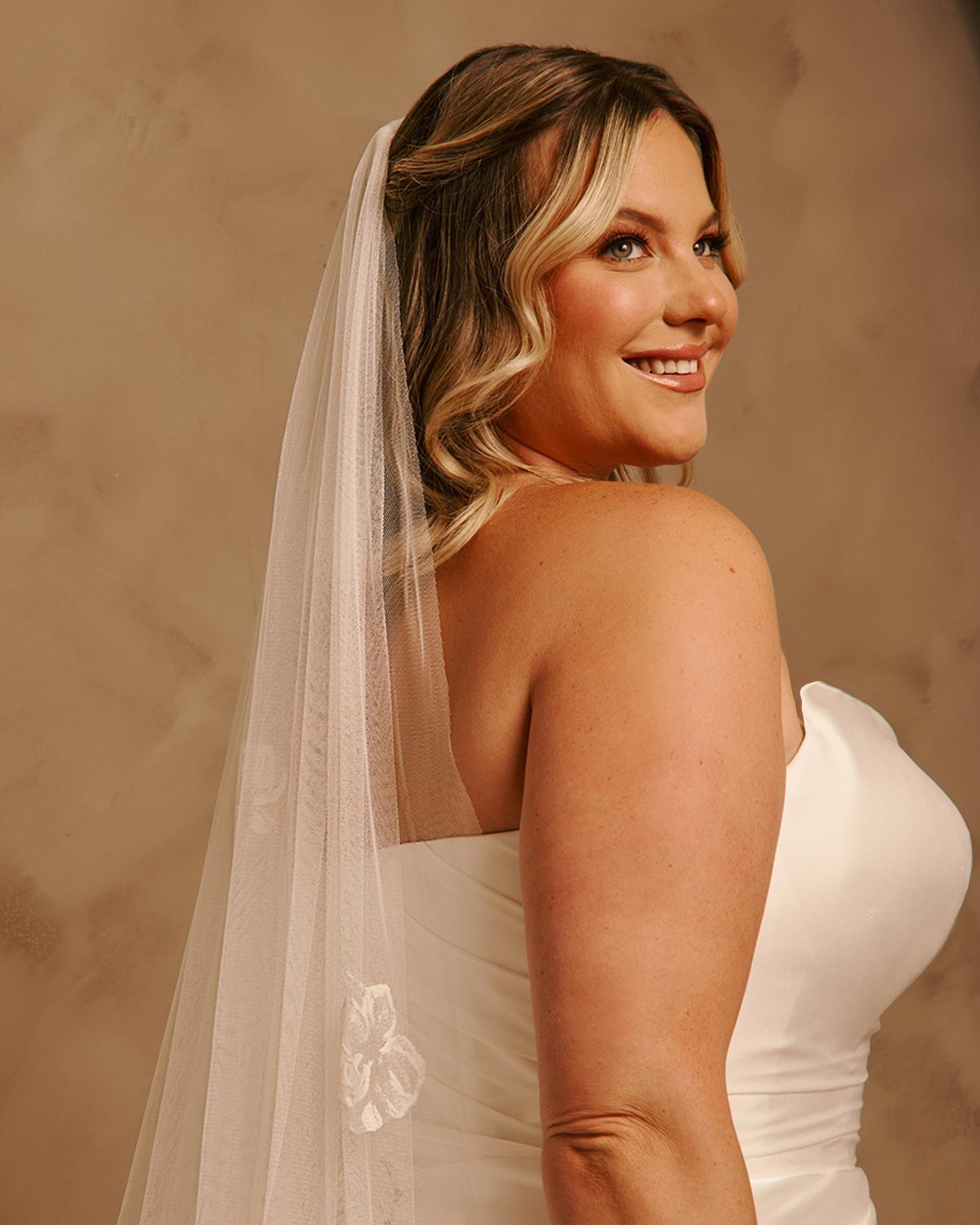 Bridal Luxe Veil with Painted Flowers Cassandra Lynne - A woman in a ivorystrapless wedding dress and long sheer veil, photographed from the back and side against a warm beige studio backdrop.