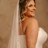 Bridal Luxe Veil with Painted Flowers Cassandra Lynne - A woman in a ivorystrapless wedding dress and long sheer veil, photographed from the back and side against a warm beige studio backdrop.