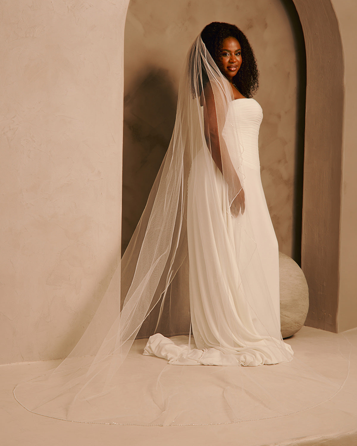 Wedding Veil with Crystal and Opaque Bead Trim A woman wearing a strapless ivory wedding gown and a long sheer tulle bridal veil, standing in a softly lit architectural setting with beige walls and arches.