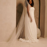 Wedding Veil with Crystal and Opaque Bead Trim A woman wearing a strapless ivory wedding gown and a long sheer tulle bridal veil, standing in a softly lit architectural setting with beige walls and arches.