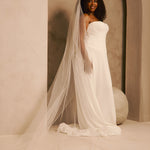 Wedding Veil with Crystal and Opaque Bead Trim A woman wearing a strapless ivory wedding gown with a long sheer veil, standing in a softly lit architectural setting with beige walls and arches.