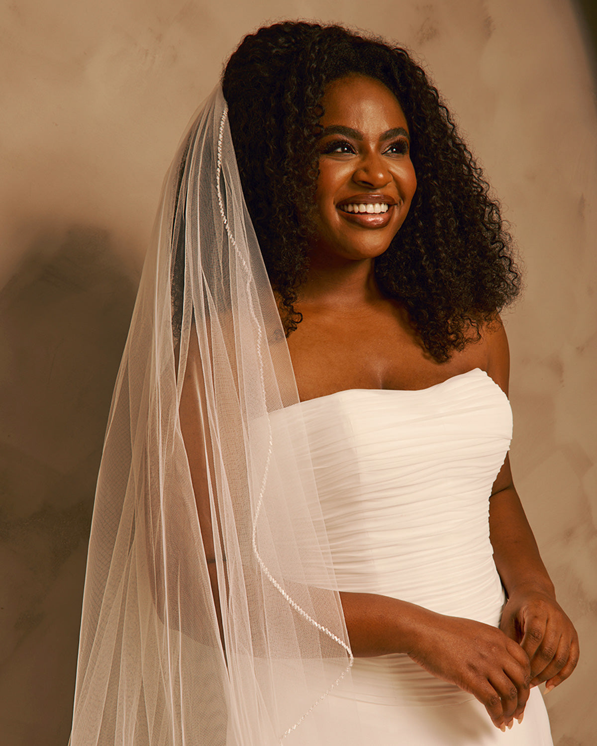 Wedding Veil with Crystal and Opaque Bead Trim A smiling woman with curly dark hair wears a strapless ivory wedding dress and a long sheer veil, posed against a warm beige studio background.
