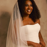 Wedding Veil with Crystal and Opaque Bead Trim A smiling woman with curly dark hair wears a strapless ivory wedding dress and a long sheer veil, posed against a warm beige studio background.