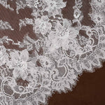 Light ivory floral lace fabric with intricate embroidered flowers and scalloped edges, laid flat against a dark brown background.