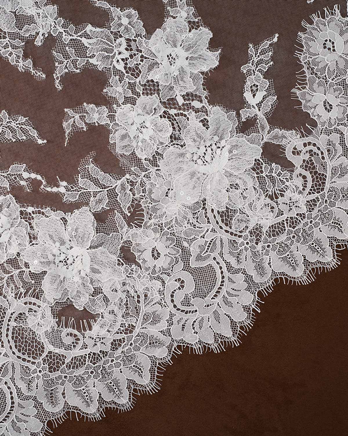 Light ivory floral lace fabric with intricate embroidered flowers and scalloped edges, laid flat against a dark brown background.