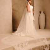 A woman wearing a long ivory wedding dress with a sheer veil stands on a stone platform in a warm beige interior with arches and a large vase in the background.