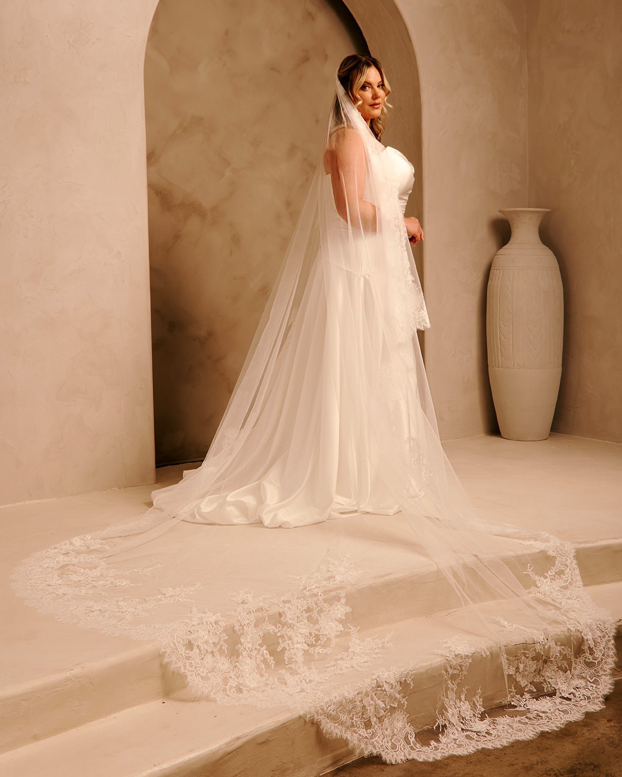 A woman wearing a long ivory wedding dress with a sheer veil stands on a stone platform in a warm beige interior with arches and a large vase in the background.
