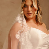 A woman wearing an ivory bridal veil with ornate floral lace trim over a white one-shoulder wedding dress, posed against a warm beige studio background.
