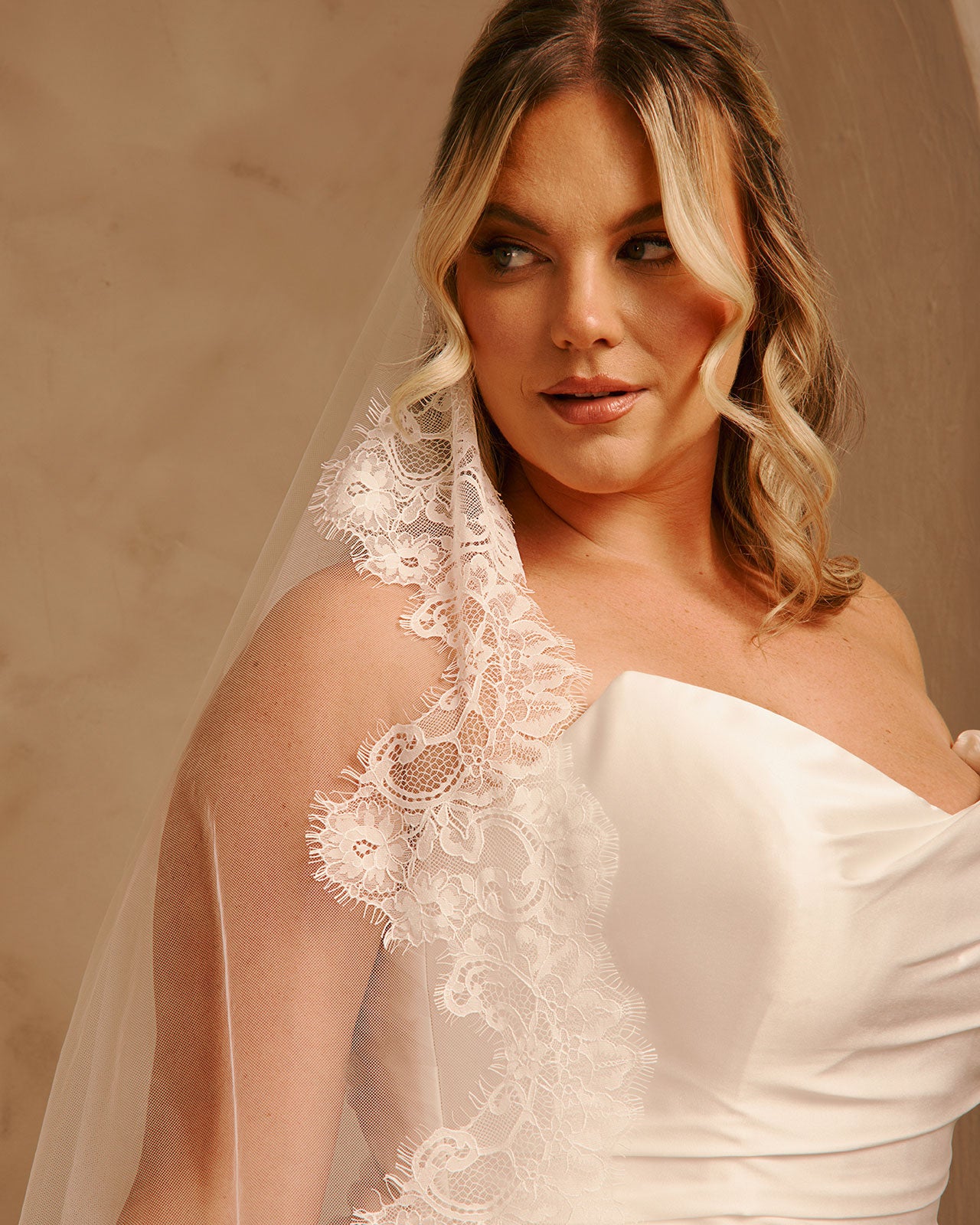 A woman wearing an ivory bridal veil with ornate floral lace trim over a white one-shoulder wedding dress, posed against a warm beige studio background.