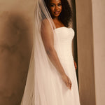A woman wearing a strapless ivory wedding gown with a long sheer veil, standing in a softly lit indoor setting with warm beige walls.