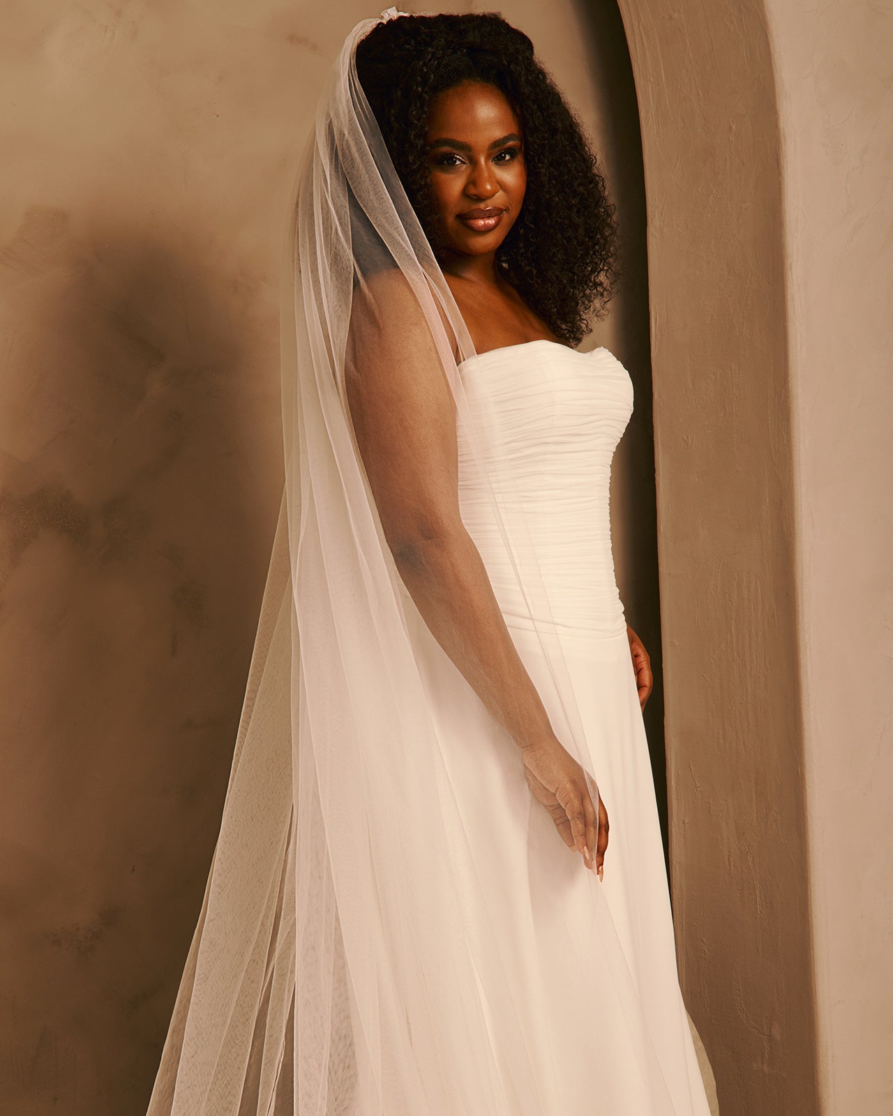 A woman wearing a strapless ivory wedding gown with a long sheer veil, standing in a softly lit indoor setting with warm beige walls.