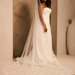 A woman wearing a strapless ivory wedding gown with a long sheer veil and floral lace detailing, standing in a warm beige interior setting.