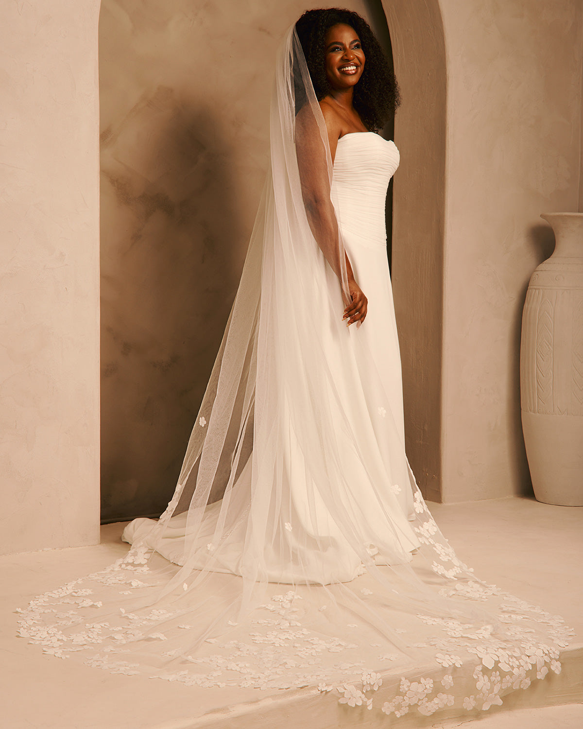 A woman wearing a strapless ivory wedding gown with a long sheer veil and floral lace detailing, standing in a warm beige interior setting.