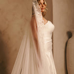 Wide Mantilla Cathedral Veil with Floral Lace Cassandra Lynne - A woman wearing a  wedding dress and a long sheer lace veil stands indoors against a warm beige wall. The veil has floral lace detailing near the top and is attached to a headpiece.