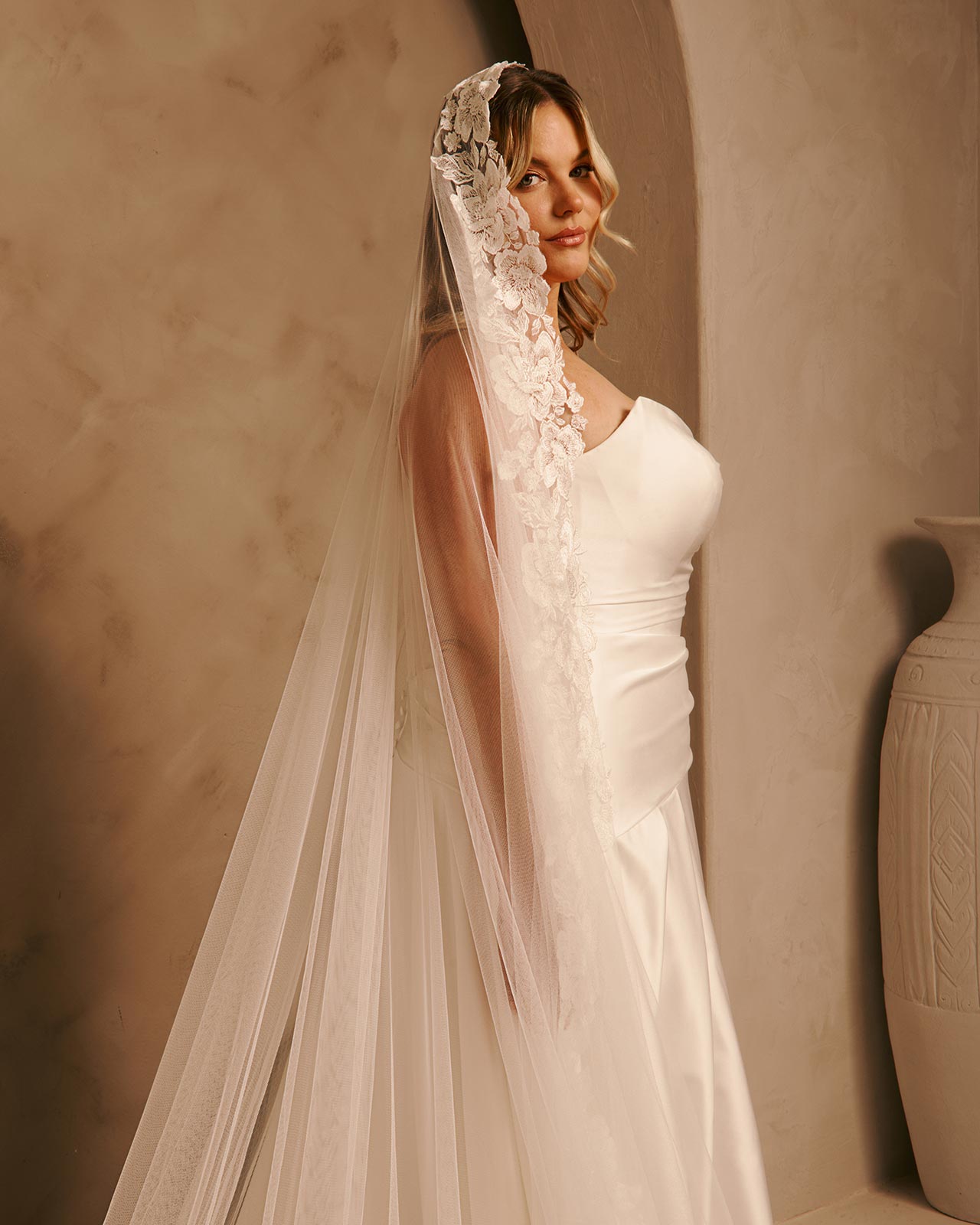Wide Mantilla Cathedral Veil with Floral Lace Cassandra Lynne - A woman wearing a  wedding dress and a long sheer lace veil stands indoors against a warm beige wall. The veil has floral lace detailing near the top and is attached to a headpiece.