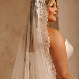 Wide Mantilla Cathedral Veil with Floral Lace Cassandra Lynne - A woman in a  bridal gown shown from the back, wearing a long sheer tulle veil with a wide floral lace border. The background is a softly lit beige wall.