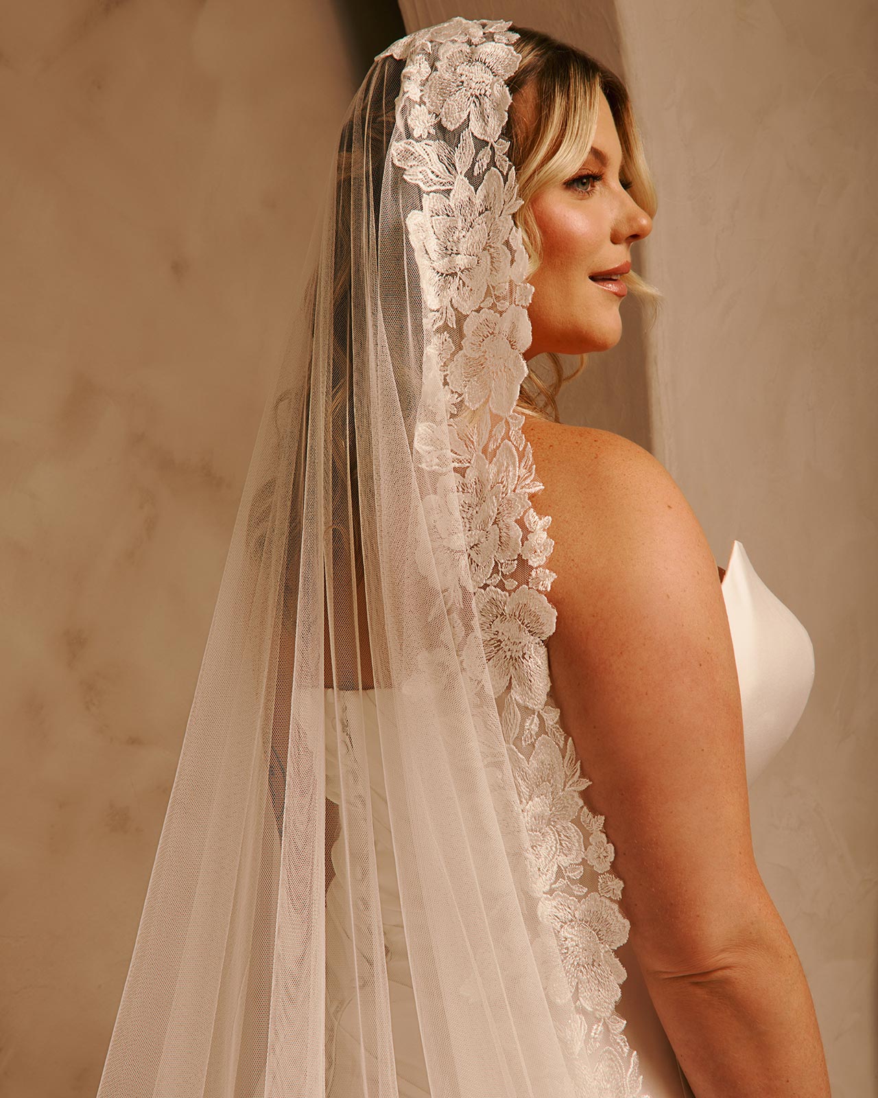 Wide Mantilla Cathedral Veil with Floral Lace Cassandra Lynne - A woman in a  bridal gown shown from the back, wearing a long sheer tulle veil with a wide floral lace border. The background is a softly lit beige wall.
