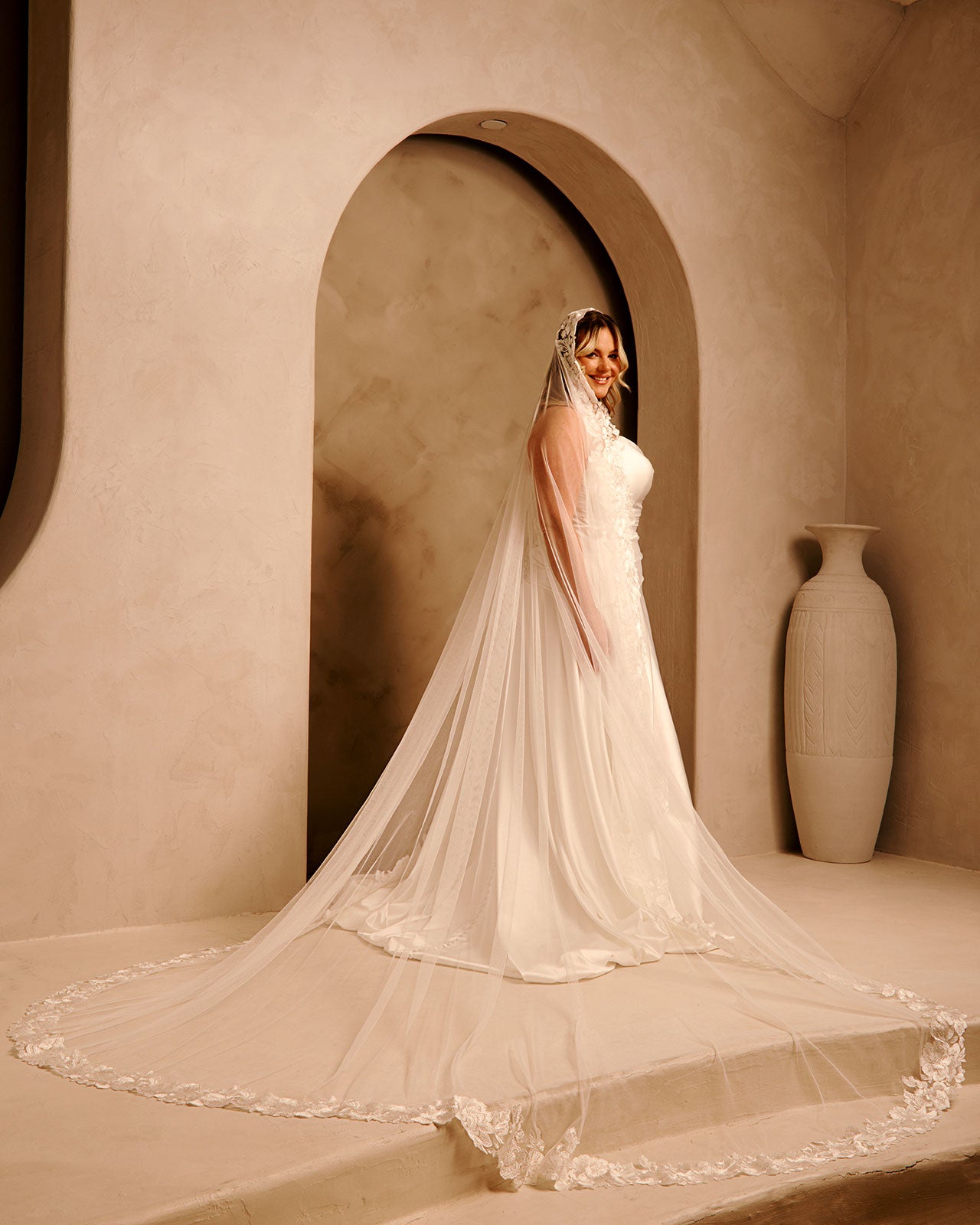 Wide Mantilla Cathedral Veil with Floral Lace Cassandra Lynne -A bride wearing a long wedding gown with a sheer veil and a very long train, standing in a warm beige architectural setting with an arched doorway and a large vase in the background.