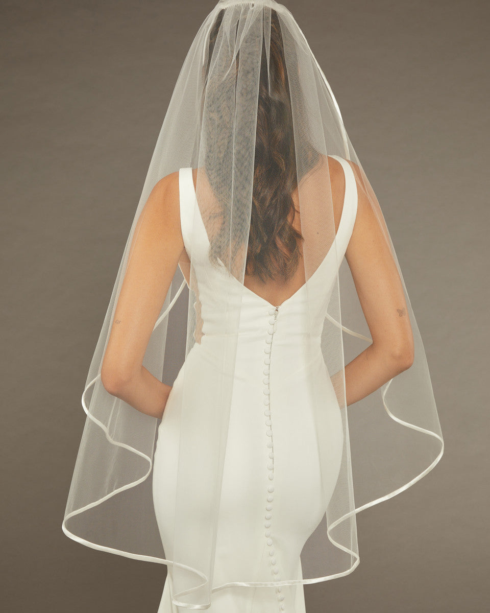 Simple Satin Trim Fingertip Bridal Veil Bride wearing a wedding dress with a sheer veil against a brown background