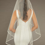 Simple Satin Trim Fingertip Bridal Veil Bride wearing a wedding dress with a sheer veil against a brown background