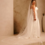 Cathedral Bridal Veil with Lace Flowers and Sequins Cassandra Lynne A woman wearing a  strapless wedding gown with a long sheer veil and lace trim, standing in a warm beige architectural setting with an archway.