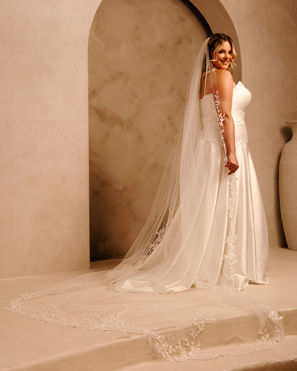 Cathedral Bridal Veil with Lace Flowers and Sequins Cassandra Lynne A woman wearing a  strapless wedding gown with a long sheer veil and lace trim, standing in a warm beige architectural setting with an archway.
