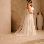 Cathedral Bridal Veil with Lace Flowers and Sequins Cassandra Lynne - A woman wearing a ivory wedding dress and long sheer veil stands in a softly lit architectural setting with beige walls, an archway, and a large vase in the background.