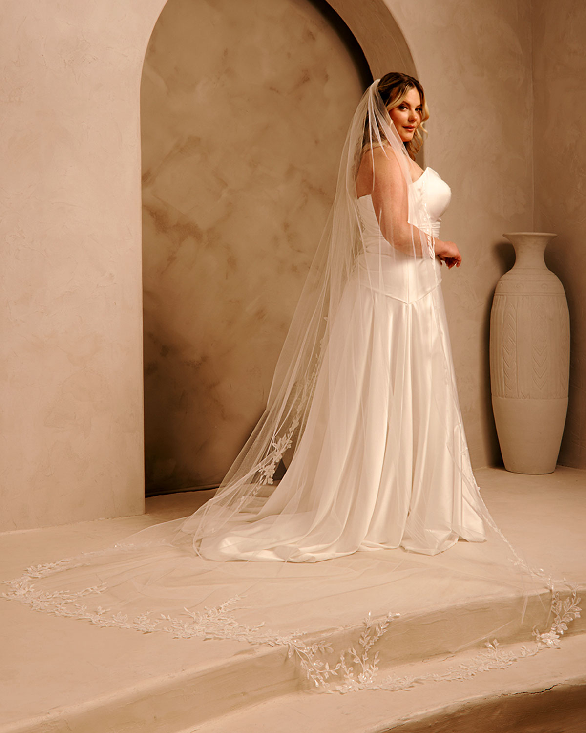 Cathedral Bridal Veil with Lace Flowers and Sequins Cassandra Lynne - A woman wearing a ivory wedding dress and long sheer veil stands in a softly lit architectural setting with beige walls, an archway, and a large vase in the background.