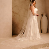Cathedral Bridal Veil with Lace Flowers and Sequins Cassandra Lynne - A woman wearing a ivory wedding dress and long sheer veil stands in a softly lit architectural setting with beige walls, an archway, and a large vase in the background.
