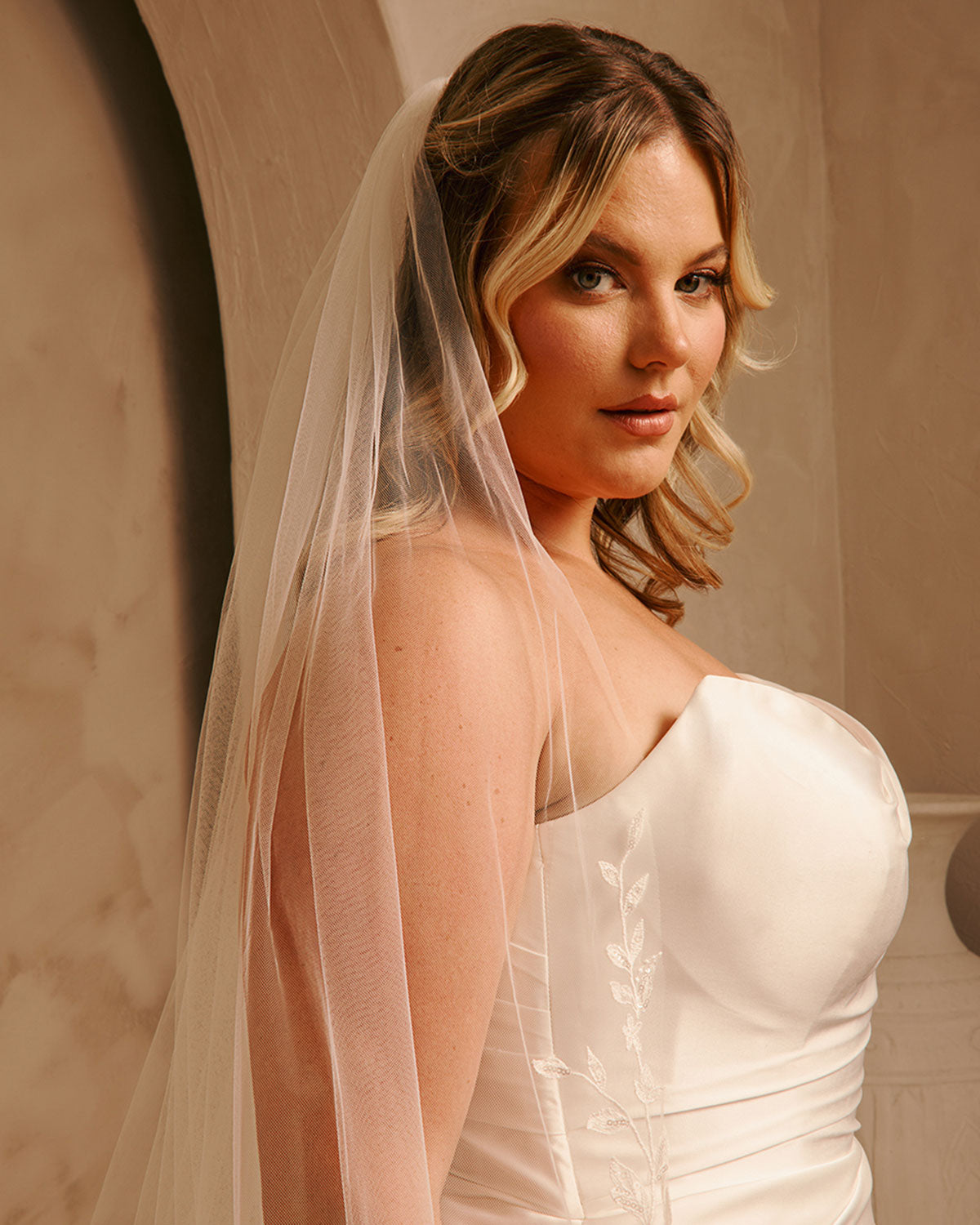 Cathedral Bridal Veil with Lace Flowers and Sequins Cassandra Lynne A woman wearing a strapless wedding dress with delicate floral embroidery and a sheer white veil, posed against a warm beige background.