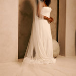 Bridal Veil with Double Crystal Trim A woman wearing a strapless ivory wedding gown with a long sheer veil, standing in a softly lit architectural setting with warm beige walls and arches.