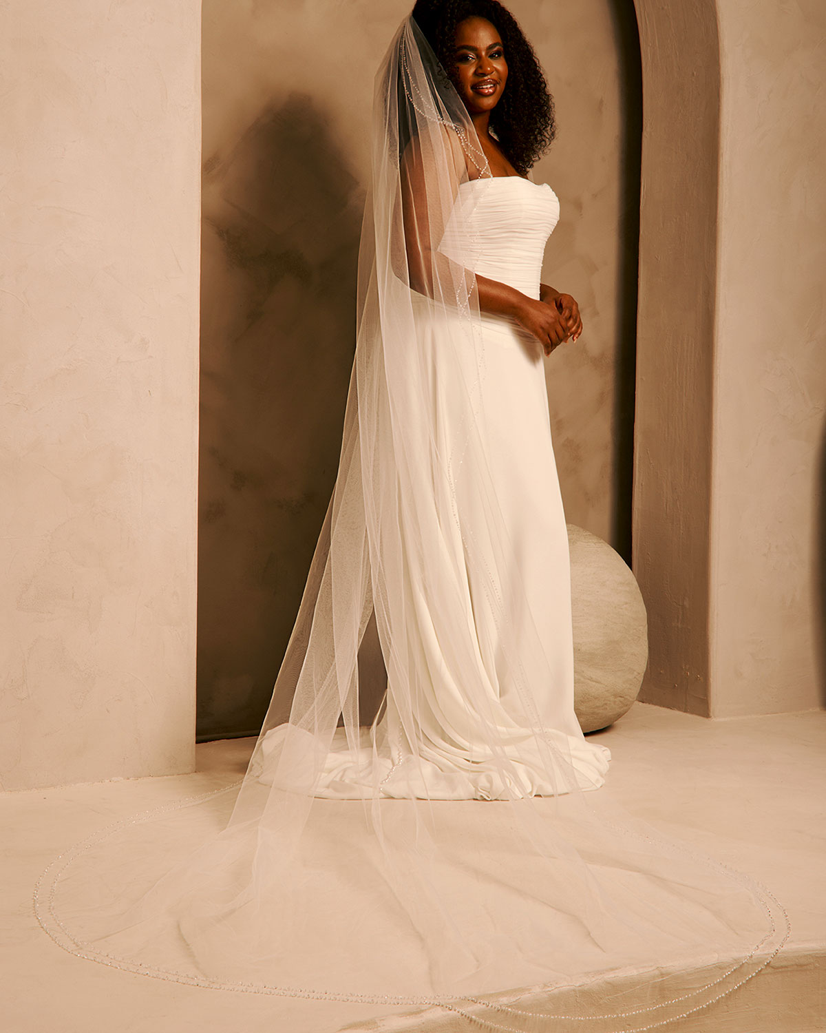 Bridal Veil with Double Crystal Trim A woman wearing a strapless ivory wedding gown with a long sheer veil, standing in a softly lit architectural setting with warm beige walls and arches.