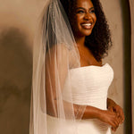 Bridal Veil with Double Crystal Trim A smiling woman with curly dark hair wears a strapless ivory wedding gown and a long sheer veil with bead detailing, posed against a warm beige wall.