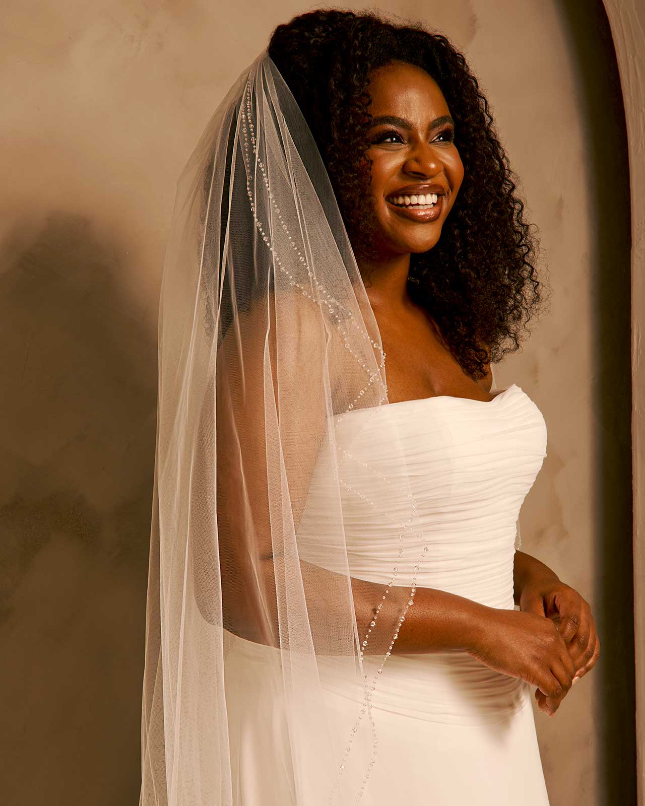 Bridal Veil with Double Crystal Trim A smiling woman with curly dark hair wears a strapless ivory wedding gown and a long sheer veil with bead detailing, posed against a warm beige wall.
