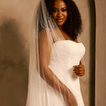 Bridal Veil with Double Crystal Trim A smiling woman wearing a strapless ivory wedding gown and a long sheer veil, posed against a warm beige studio backdrop.