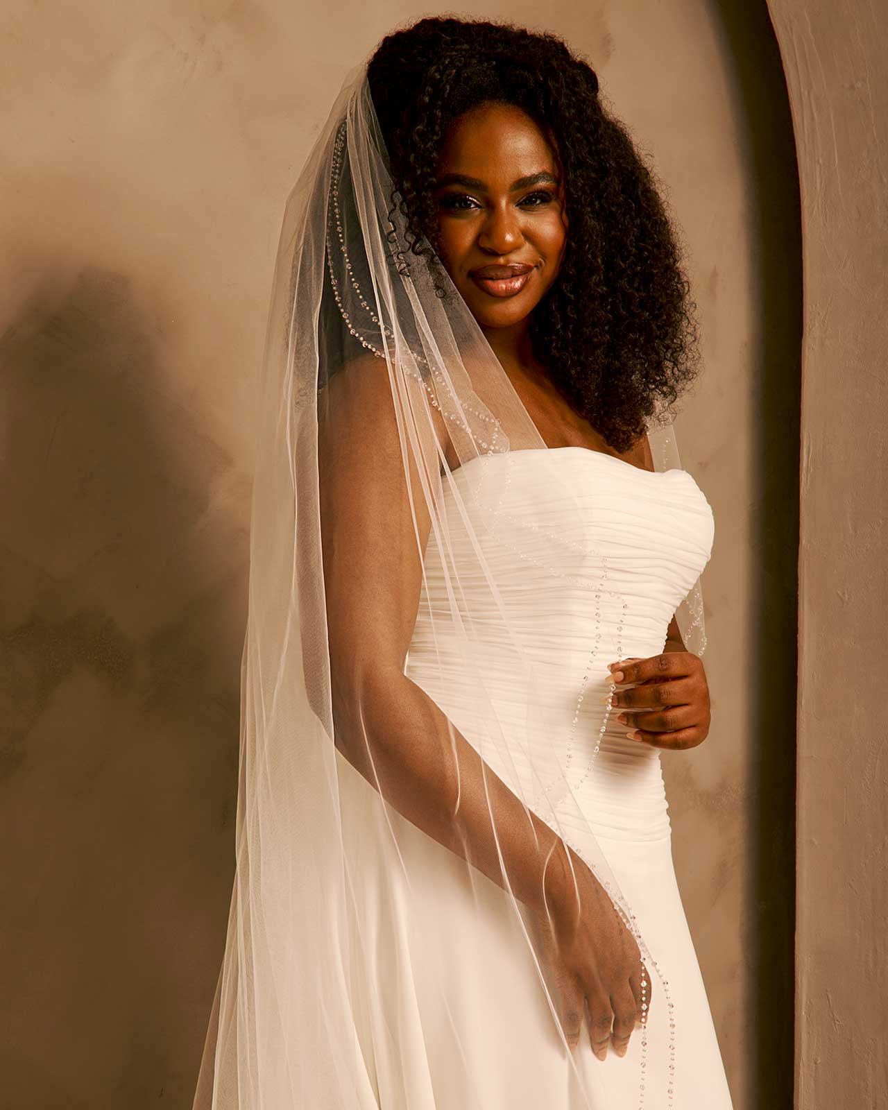 Bridal Veil with Double Crystal Trim A smiling woman wearing a strapless ivory wedding gown and a long sheer veil, posed against a warm beige studio backdrop.