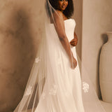 Bridal Veil with Painted Flowers and Pearls Cassandra Lynne - A woman wearing a strapless ivory wedding gown and a long sheer tulle bridal veil with white floral embroidery, standing in a warm beige studio setting with a large vase at the side.