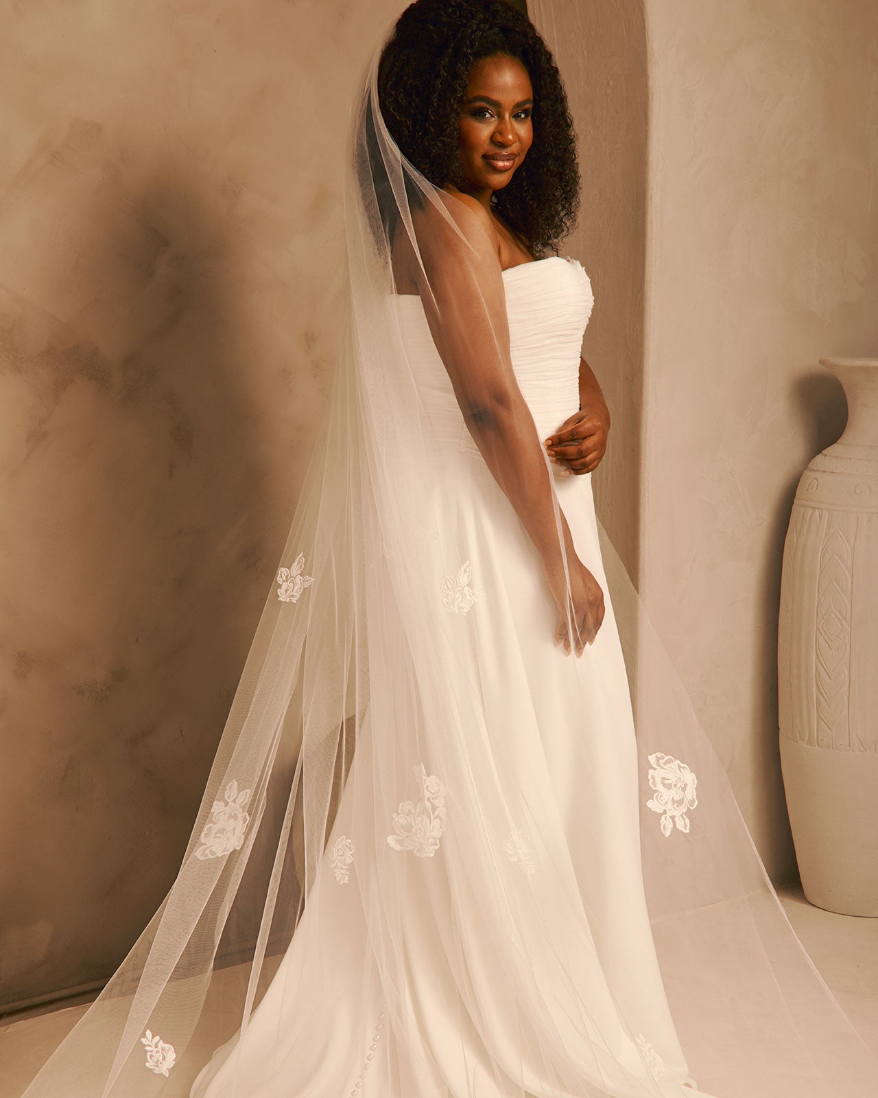 Bridal Veil with Painted Flowers and Pearls Cassandra Lynne - A woman wearing a strapless ivory wedding gown and a long sheer tulle bridal veil with white floral embroidery, standing in a warm beige studio setting with a large vase at the side.