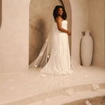 Bridal Veil with Painted Flowers and Pearls Cassandra Lynne - A woman wearing a strapless ivory wedding gown with a long sheer veil decorated with floral appliqués, standing in a warm beige architectural setting with an arched alcove and a large vase.