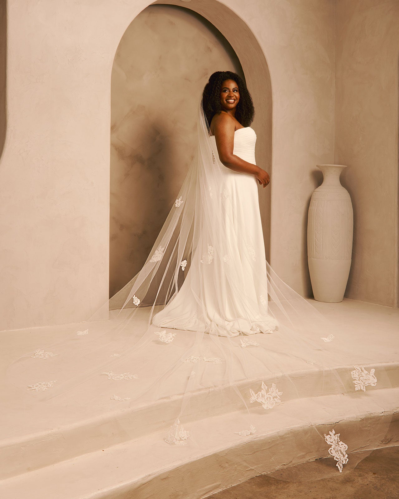 Bridal Veil with Painted Flowers and Pearls Cassandra Lynne - A woman wearing a strapless ivory wedding gown with a long sheer veil decorated with floral appliqués, standing in a warm beige architectural setting with an arched alcove and a large vase.