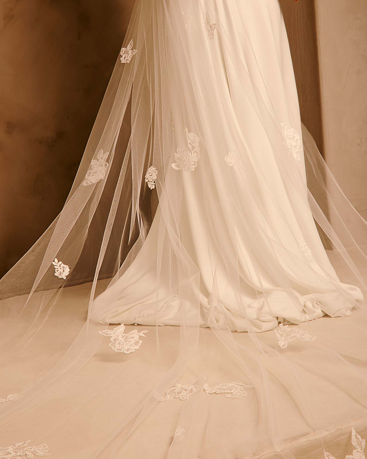 Bridal Veil with Painted Flowers and Pearls Cassandra Lynne - A close-up of a bridal veil with delicate ivory floral lace appliques, draped over a white wedding gown against a warm, neutral background.